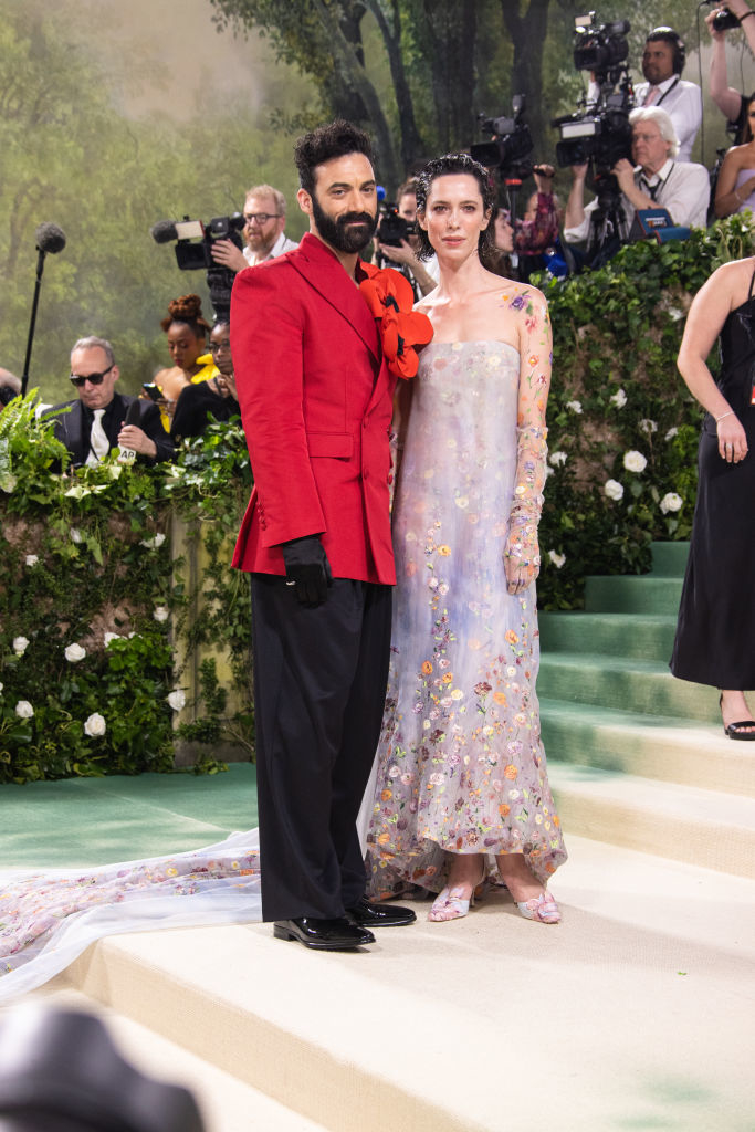 Morgan in suit with large flower, Rebecca in a floral sheer dress, photographers in background