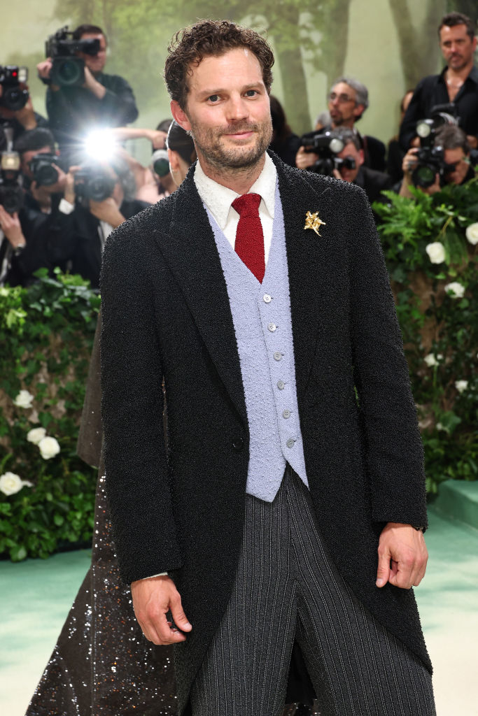 Jamie Dornan in textured coat over vest and tie, with photographers in background