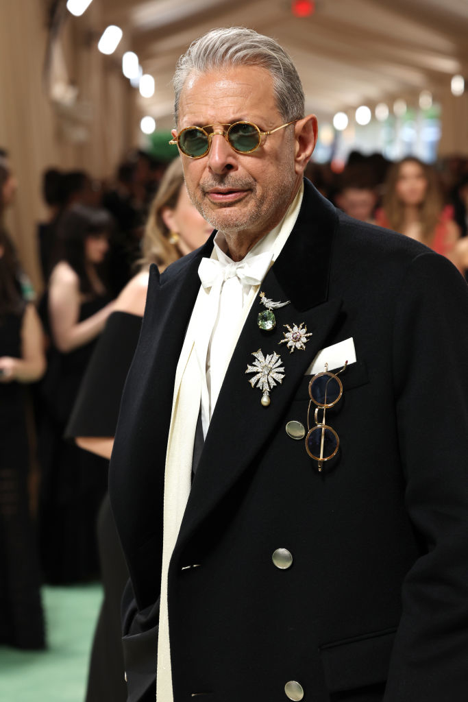 Jeff Goldblum in tuxedo with bow tie and sunglasses, adorned with star-shaped brooch and round glasses on lapel