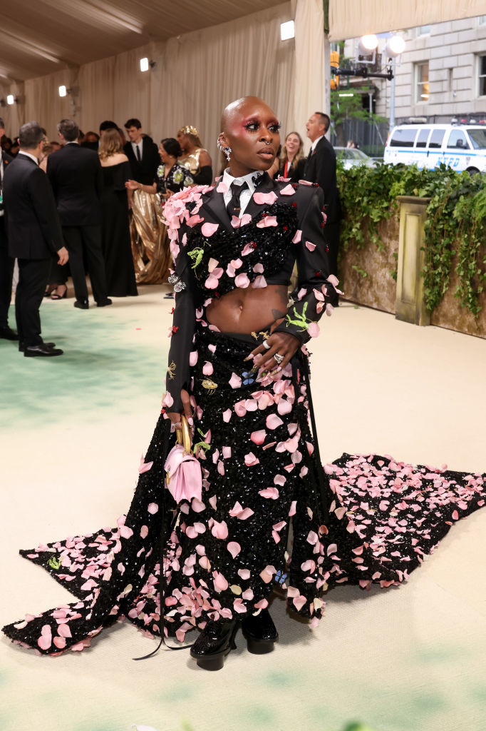 Cynthia Erivo on a red carpet wearing a black outfit adorned with pink floral motifs and cut-outs, standing confidently