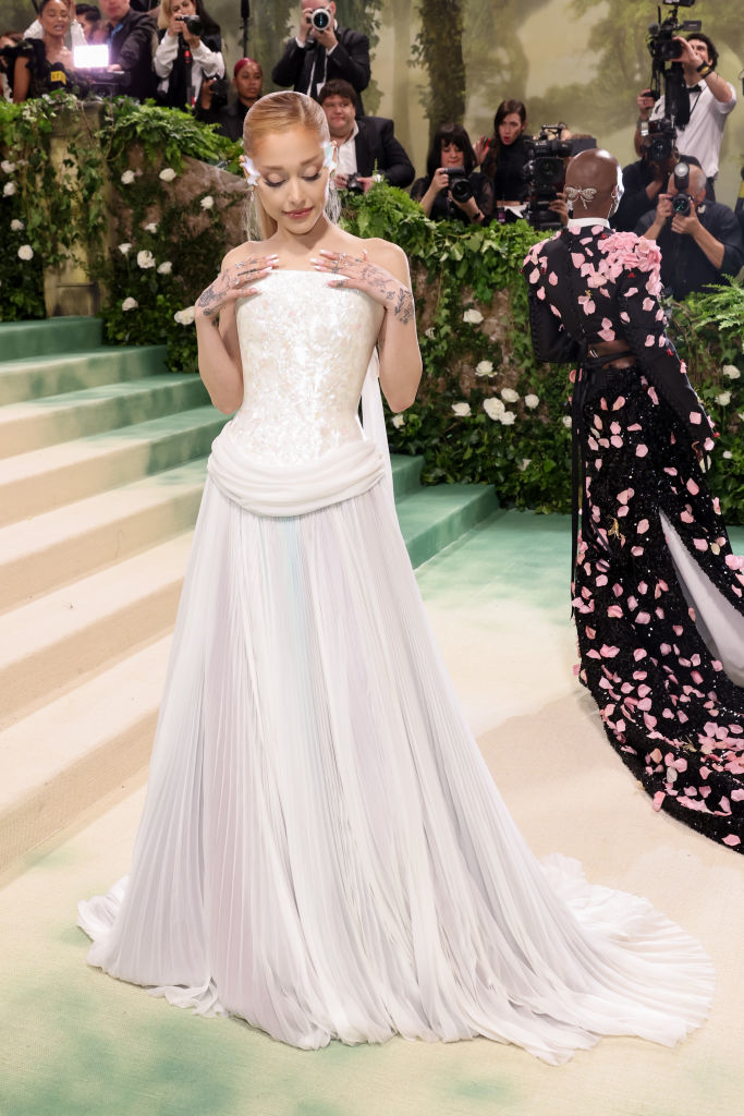 Ariana in gown with pleated skirt and embellished top is standing with hands near face, photographers in background