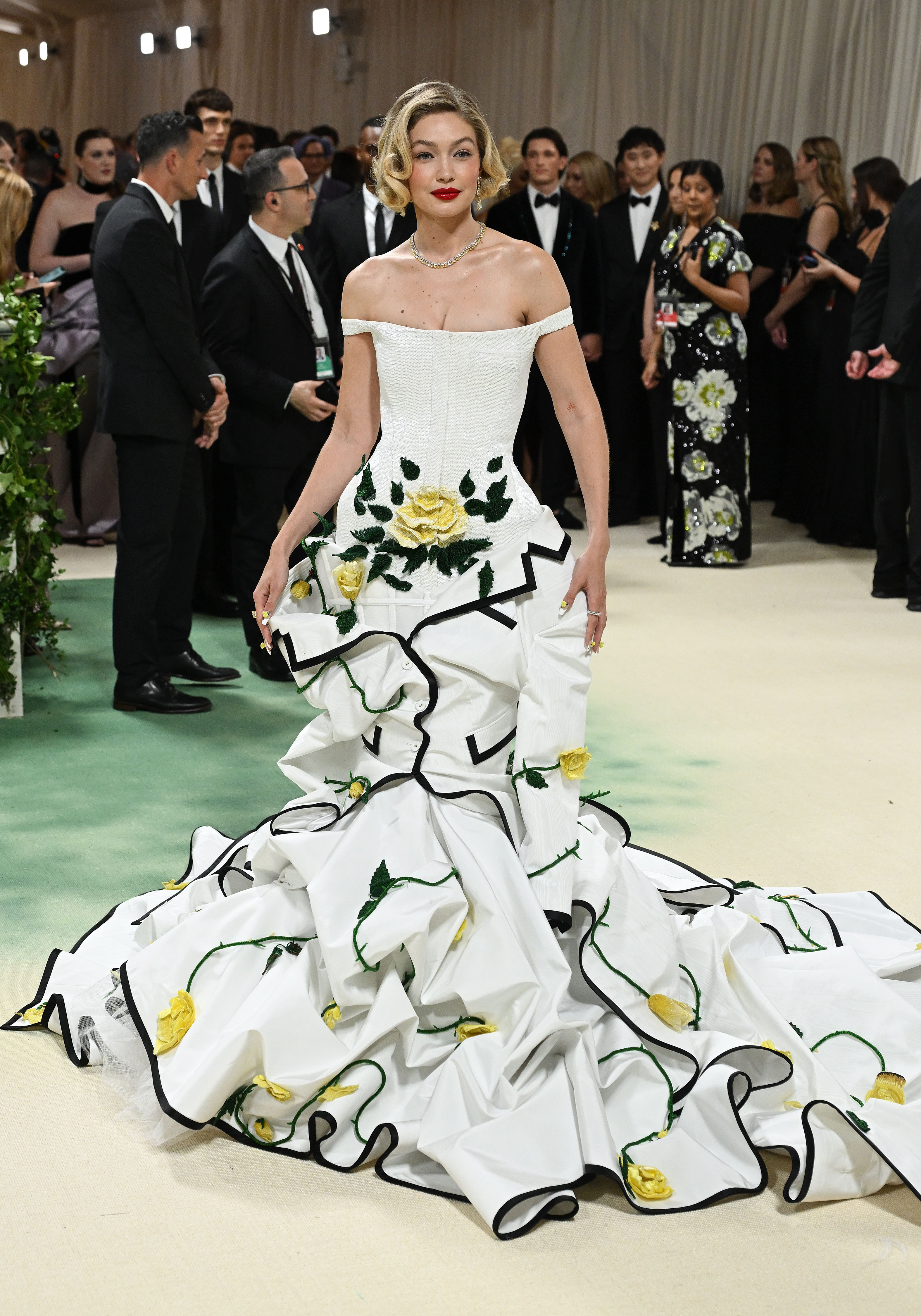 Gigi Hadid in a white cascading gown with black trim and yellow rose accents, standing at an event