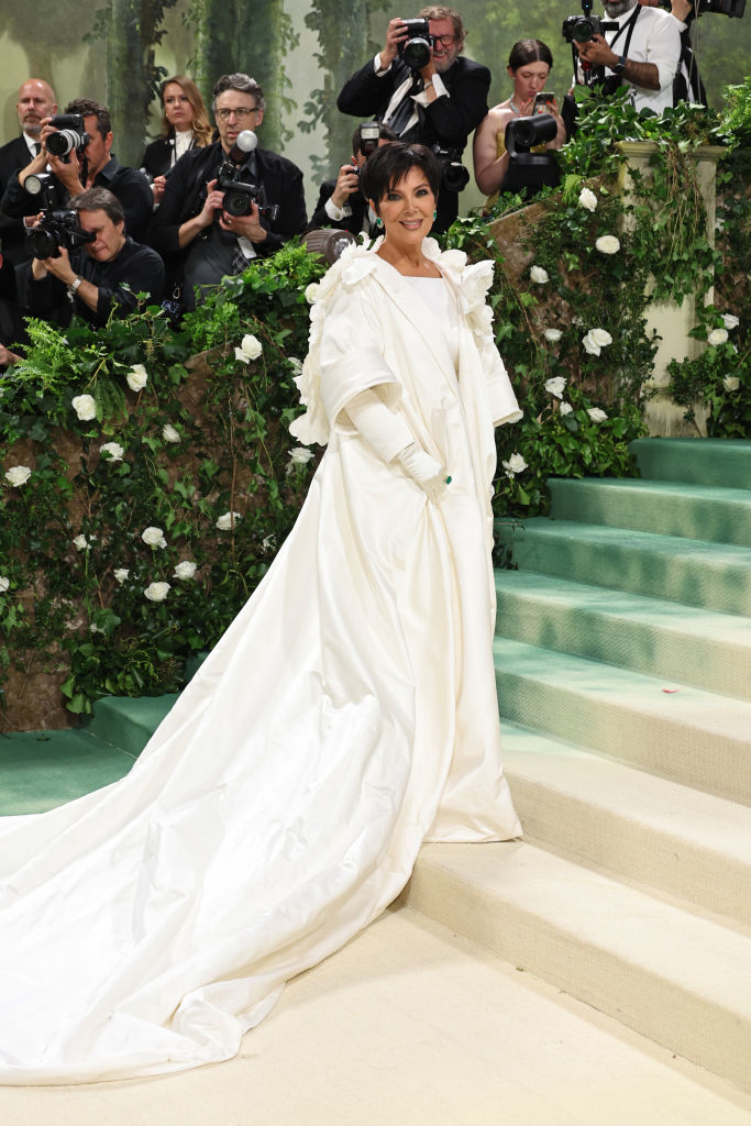 Kris Jenner in a white ruffled outfit with a long train on stairs at a gala event. Photographers in the background
