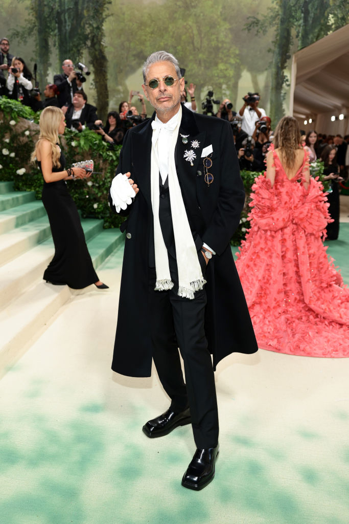 Jeff Goldblum in black tuxedo with long coat, white scarf, and sunglasses posing at an event
