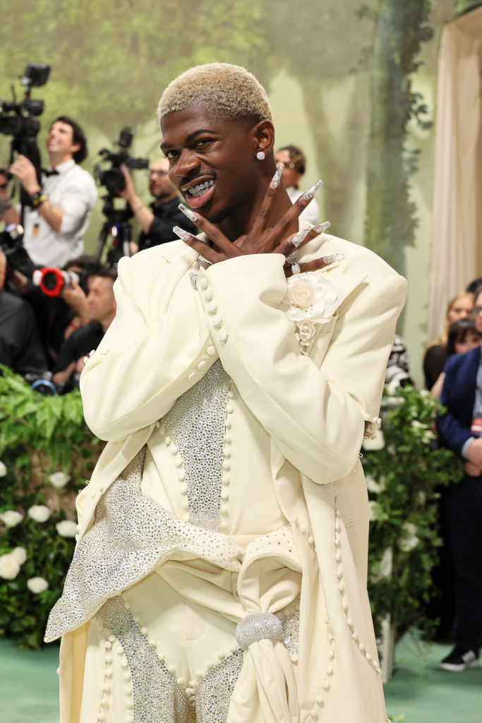 Lil Nas X in an ornate cream suit with embellishments posing with hands crossed at an event