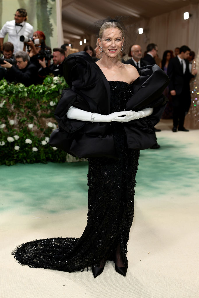 Naomi Watts in an off-shoulder black gown with puffy sleeves and sequined skirt, wearing long white gloves, on a carpeted area