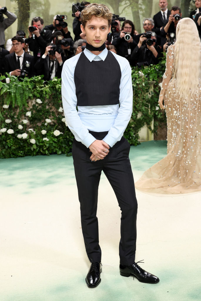 Troye Sivan in a sleeveless black vest and tie, dress shirt, trousers, and shiny shoes stands on a carpet, photographers in the background