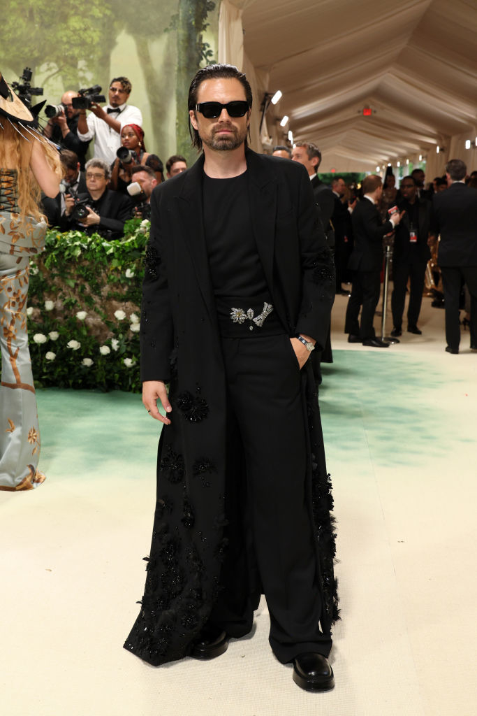 Sebastian Stan on red carpet in black outfit with embellished coat, sunglasses, and slicked-back hair