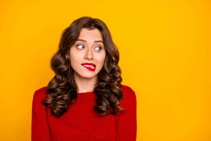 Woman with wavy hair and red lipstick looks away with a puzzled expression. She wears a red sweater