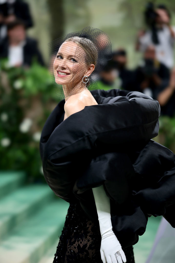 Naomi Watts in a black gown with puff sleeves and white gloves, posing at an event