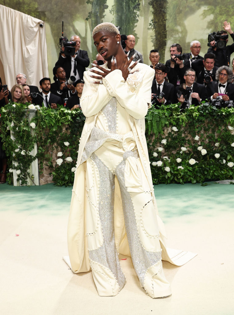 Lil Nas X posing in an embellished white suit with a draped sash at a glam event with photographers in the background