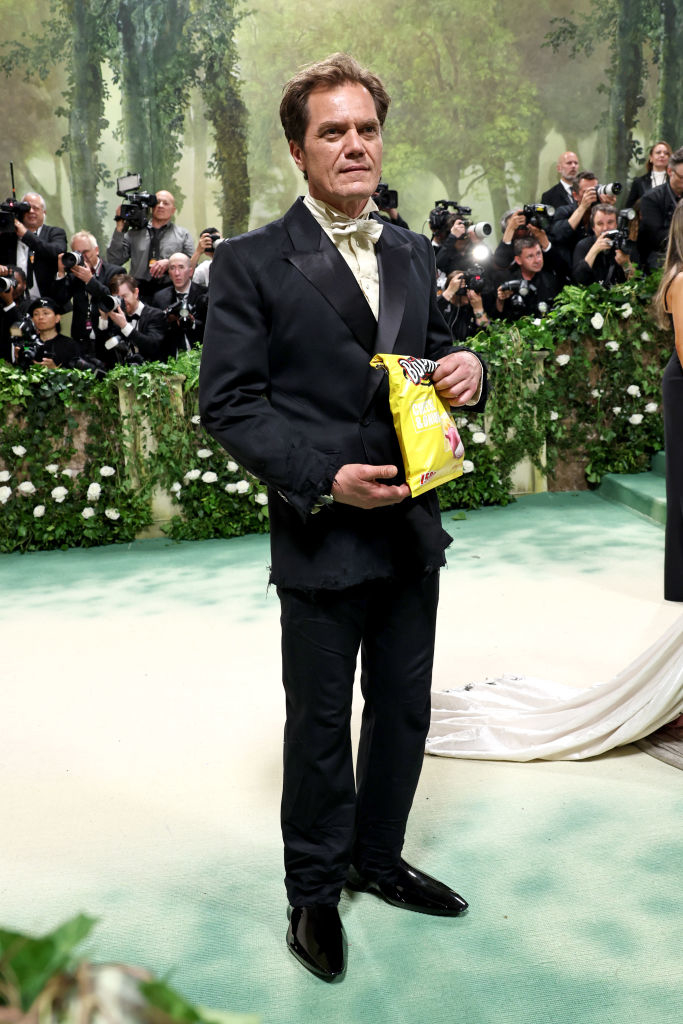 Michael Shannon in black tuxedo holding snack bag, standing in front of photographers at event