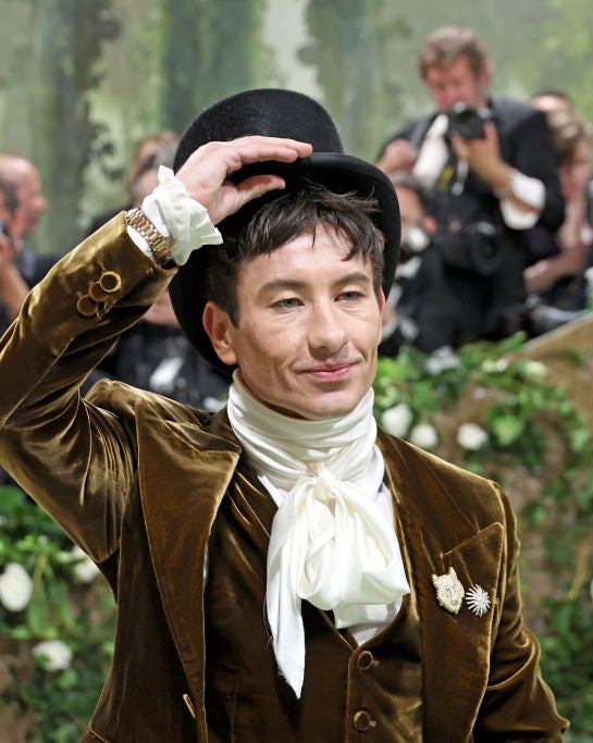 Barry Keoghan tipping hat, wearing brown jacket, white cravat, on greenery backdrop, photographers in background