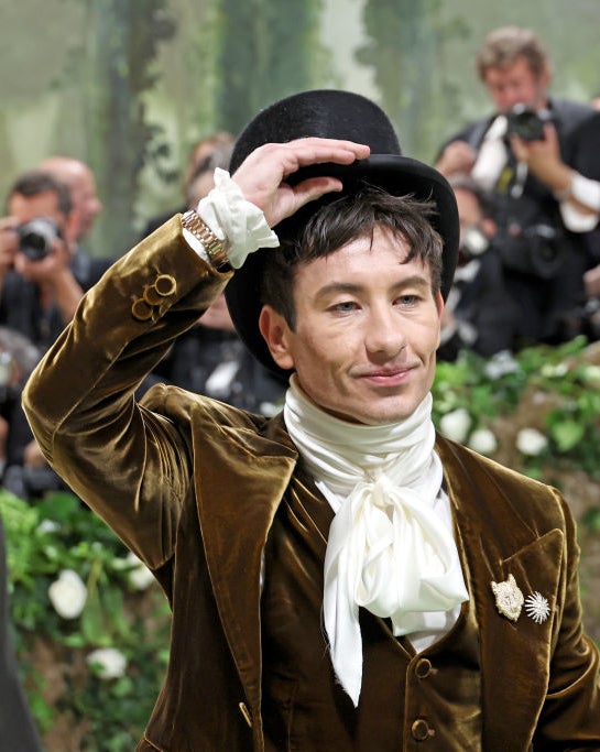 Barry Keoghan tipping hat, wearing brown jacket, white cravat, on greenery backdrop, photographers in background