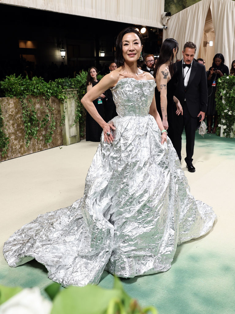 Michelle Yeoh in a metallic textured gown with a full skirt posing on the carpet, tattoos visible on her arm