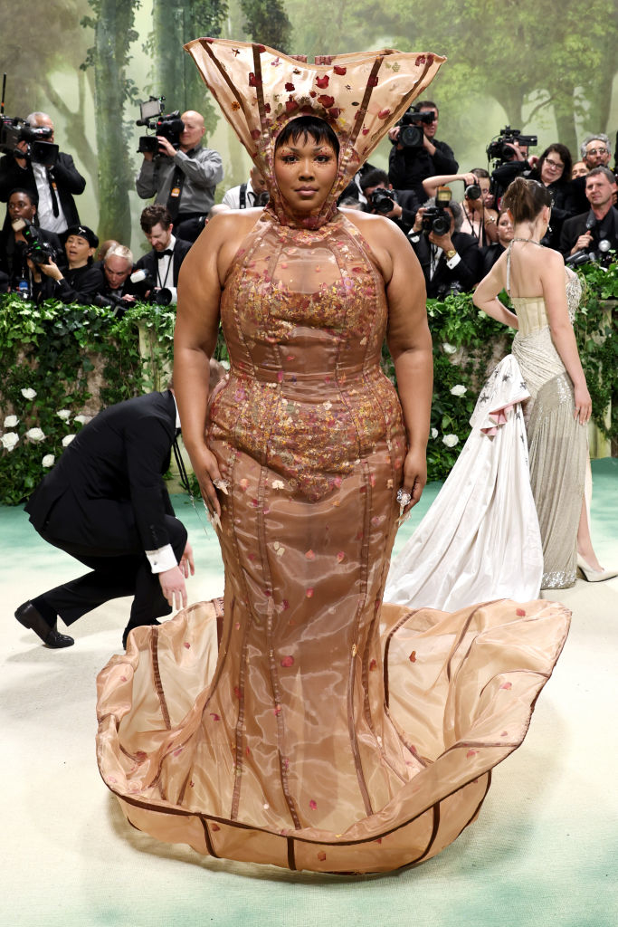 Lizzo wearing a beaded gown with a ruffled hem and a headpiece at an event with photographers in the background