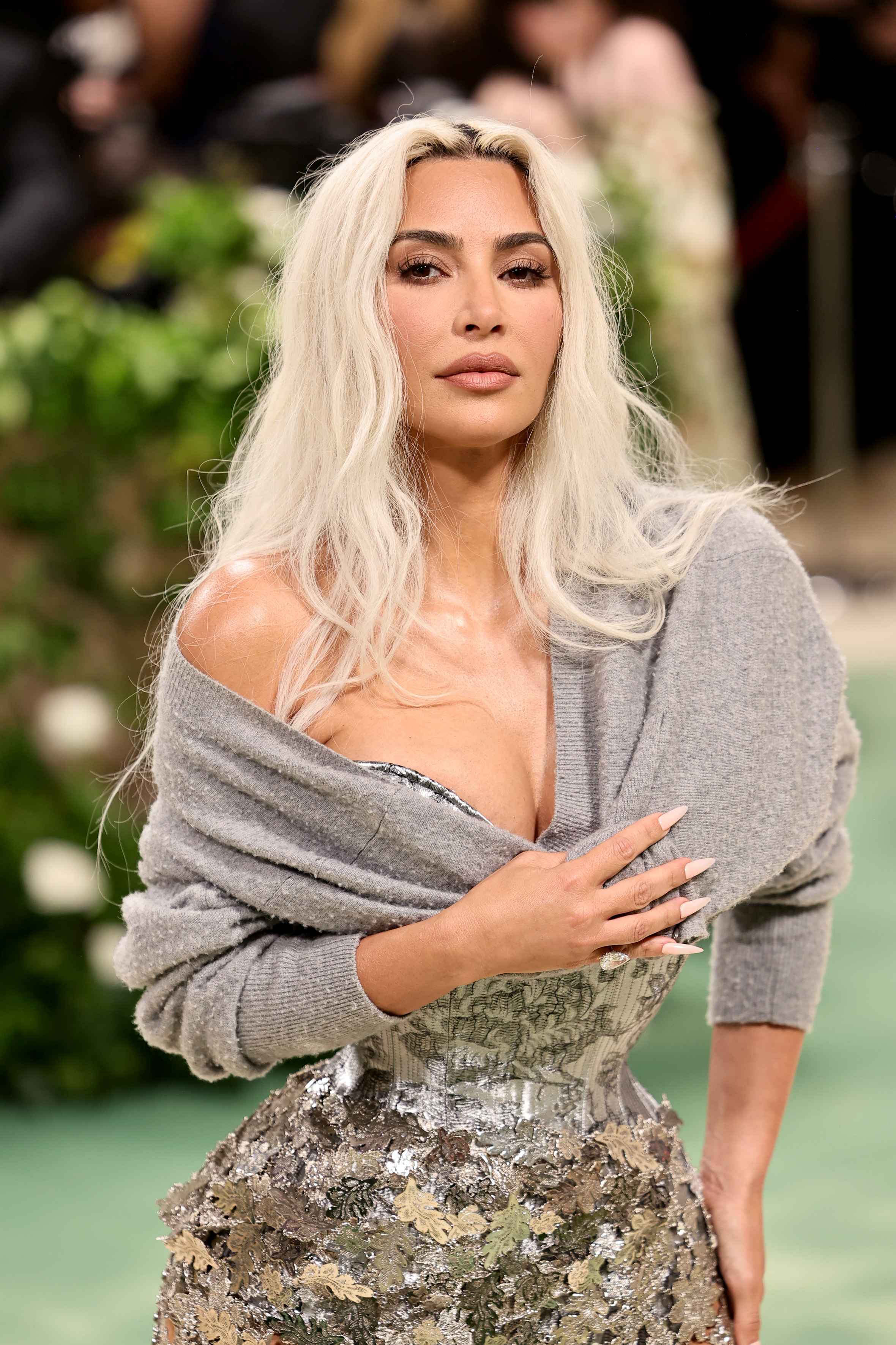 Kim Kardashian posing in a grey top with silver details, hand on chest, at an event