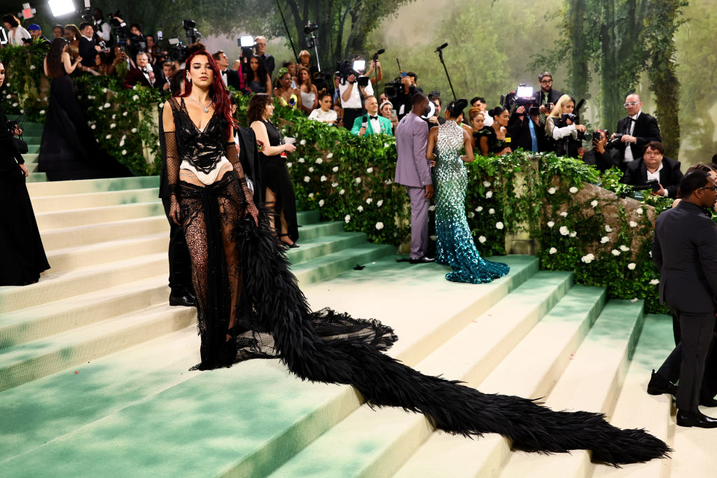 Person in a black lace outfit with a long trail standing on Met Gala stairs with attendees in background