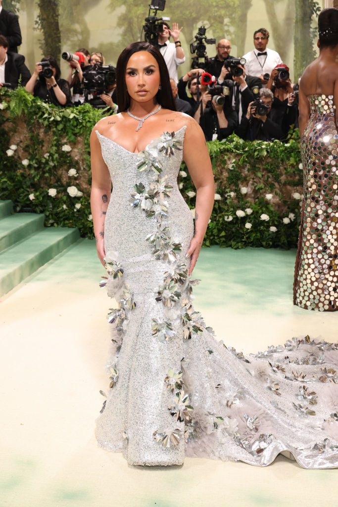 Demi Lovato in a sparkling sleeveless gown with silver floral embellishments, standing on a themed event backdrop
