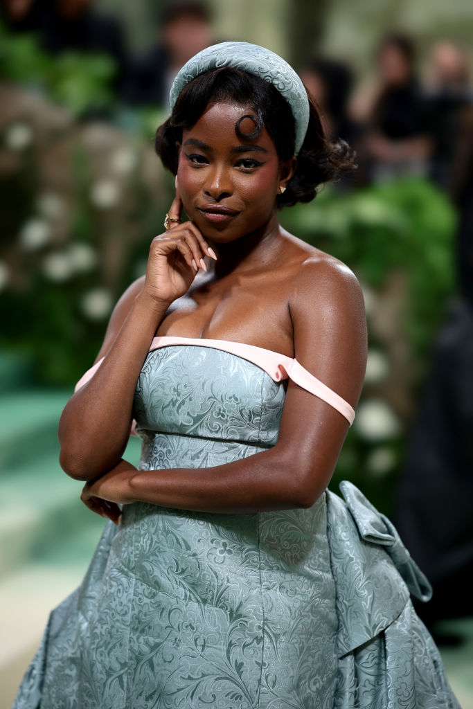 Amanda Gorman at event wearing a strapless gown with a headpiece, posing with hand on cheek