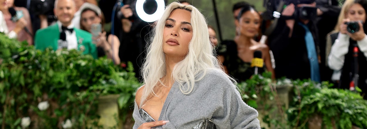 Kim Kardashian in a fitted, shimmering silver gown with a trail, posing on steps at an event with cameras in the background