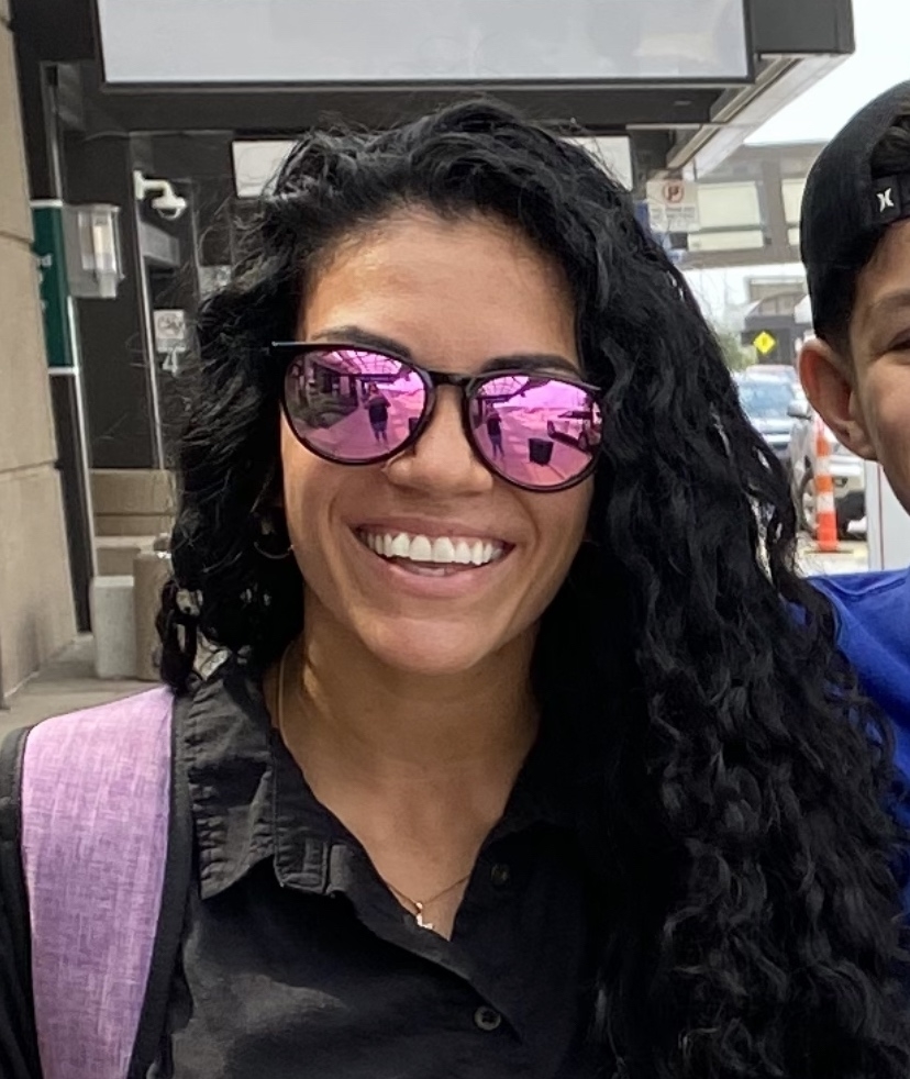 Woman in sunglasses with a partial view of a boy behind her. She smiles, wearing a casual shirt and backpack