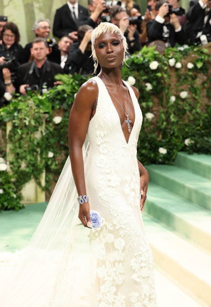 A person in an elegant white lace gown with a plunging neckline and trail, accessorized with bold jewelry, posing on stairs