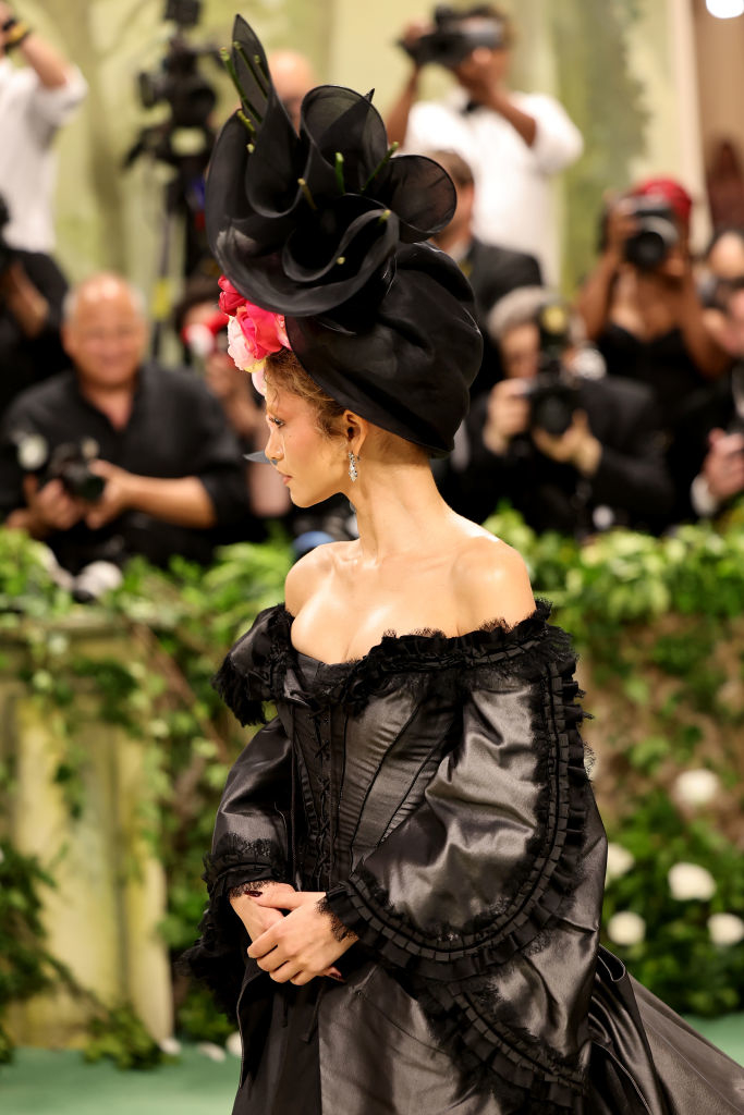 Zendaya at event wearing an elaborate hat and ruffled off-shoulder dress. Photographers in the background