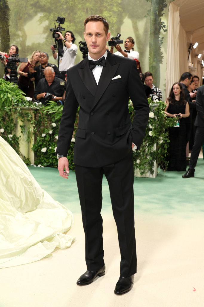 Alexander Skarsgard in a classic black tuxedo with bow tie at an event with photographers in background