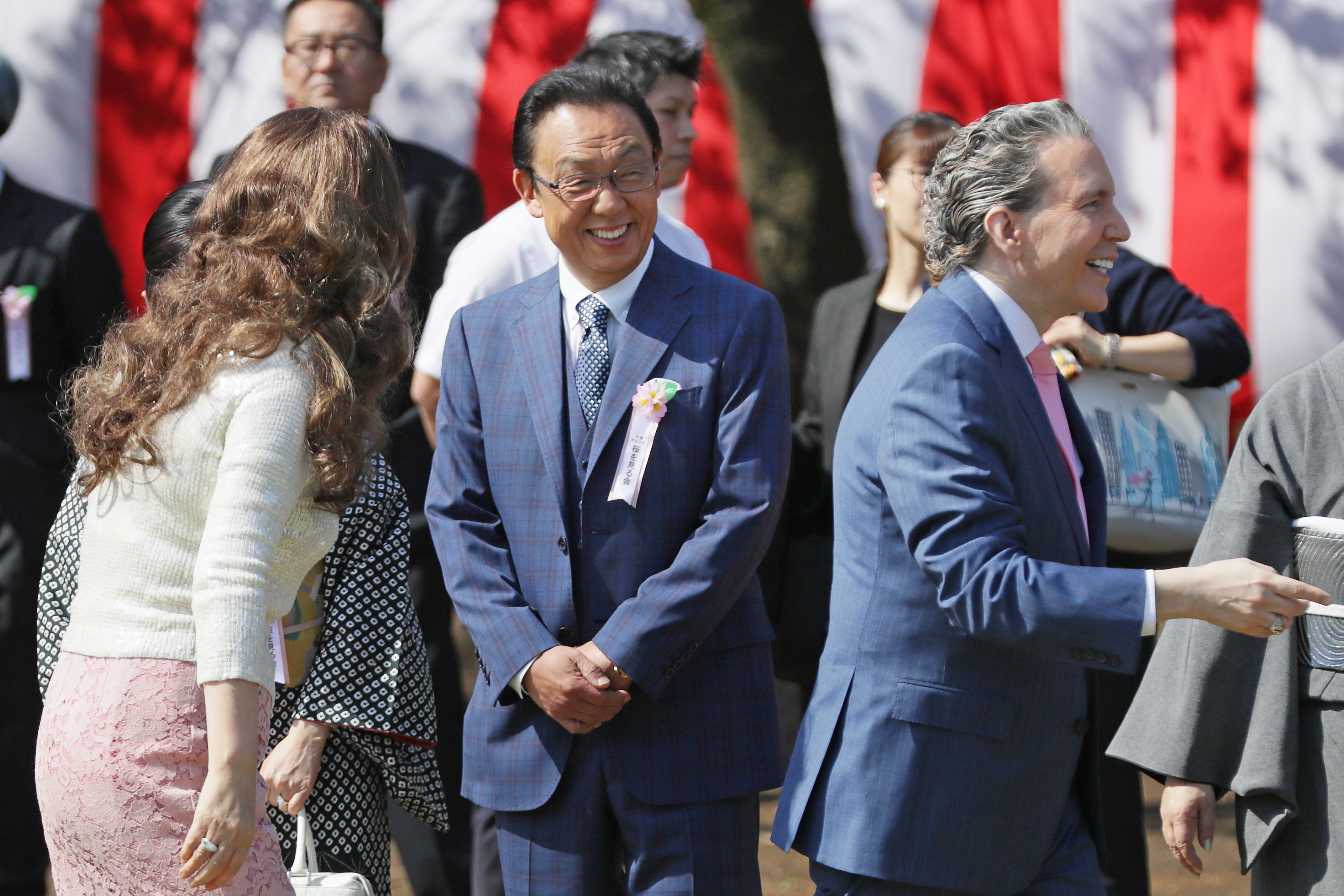 春のイベントに出席する笑顔の人々、梅沢富美男さんはスーツ着用、背景に旗