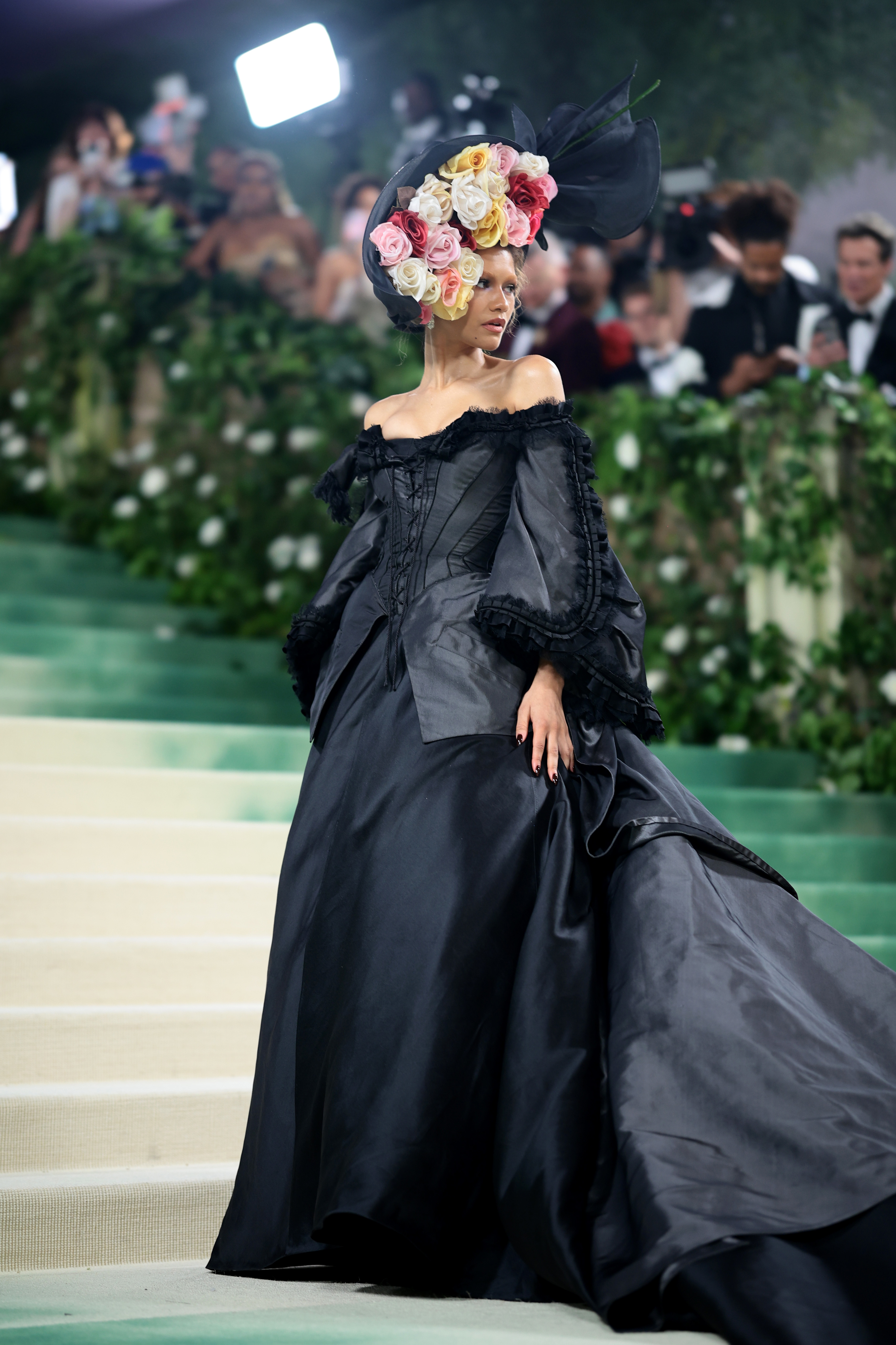 Celebridad en vestido de gala negro con sombrero grande adornado con flores desfilando en evento