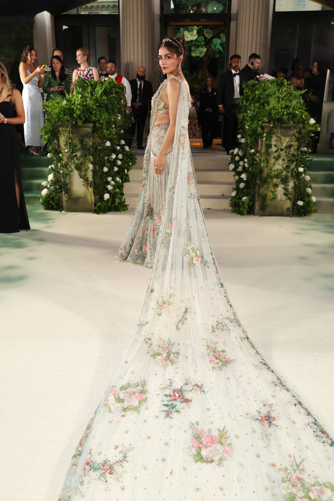 Person in an elegant floral gown with a long train, on a path bordered by greenery and spectators