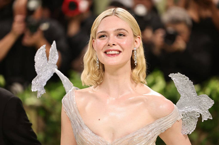 Elle Fanning smiles at an event wearing a sheer, shimmering dress with whimsical winged shoulders