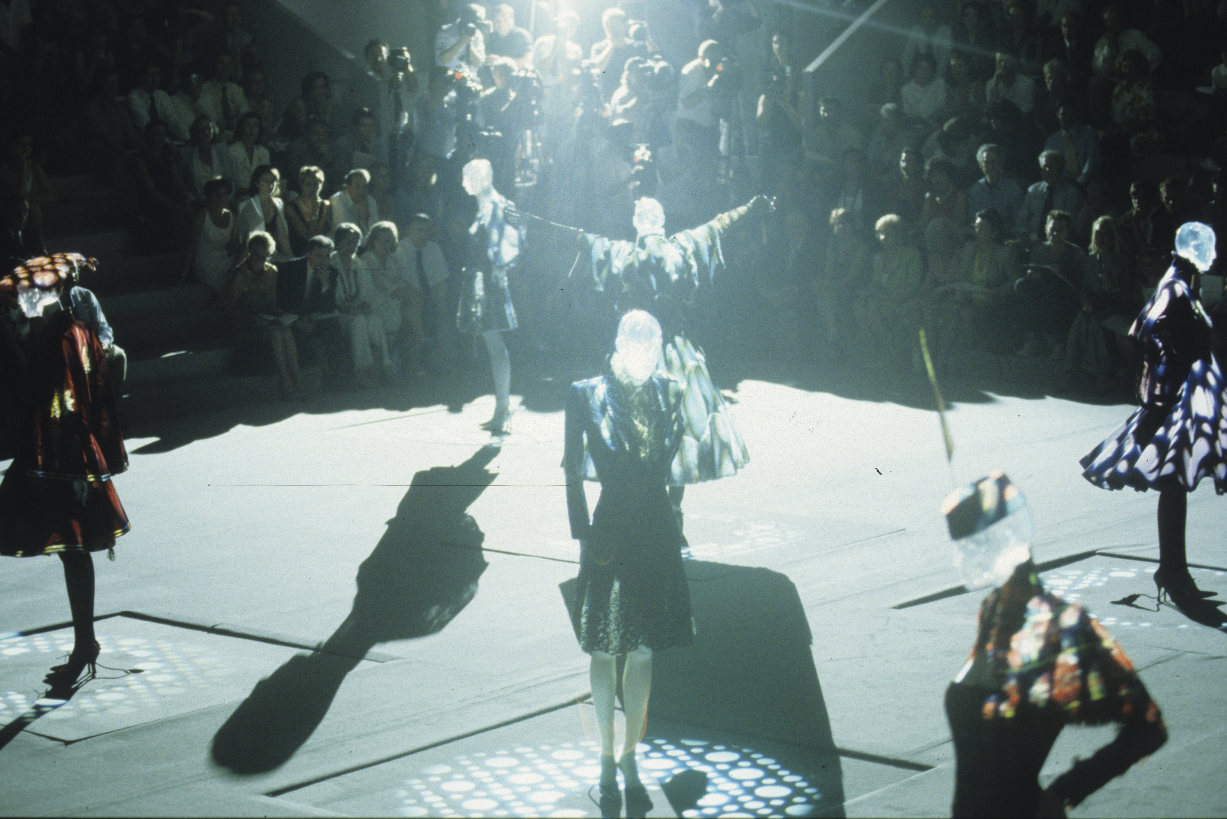 Fashion models walk a runway with patterned clothing in a spotlight, surrounded by an audience