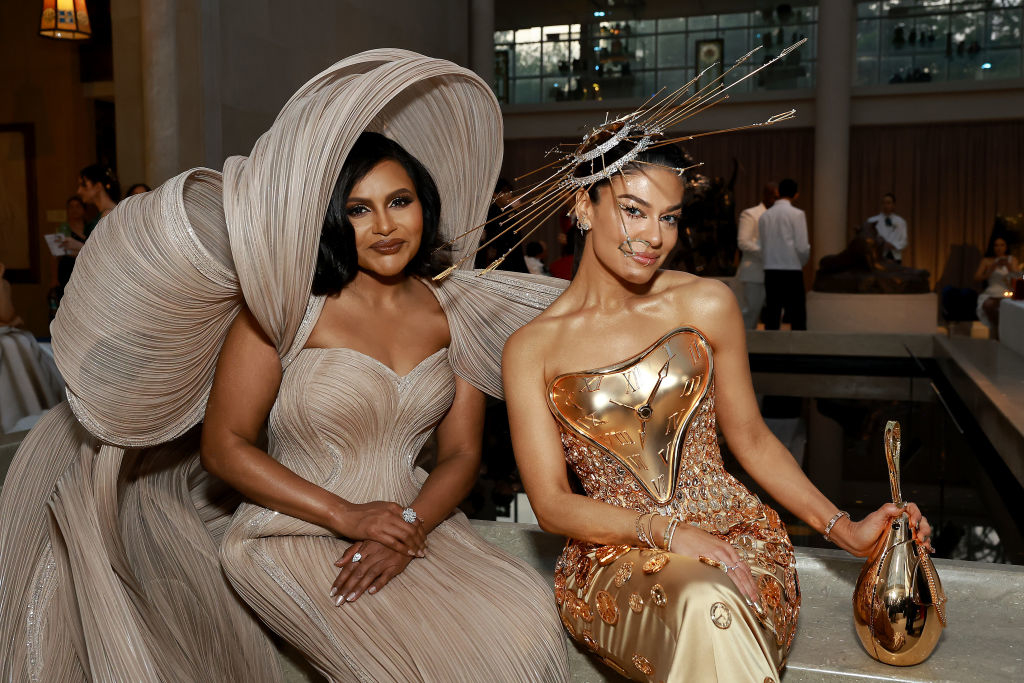 Two women at an event, one wearing a voluminous dress with a large hat, the other in a metallic, figure-hugging gown with a headpiece