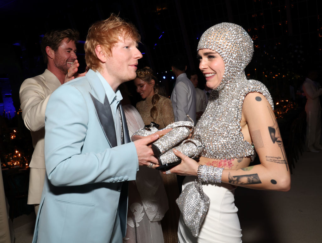 Ed Sheeran in a blue suit and a person in a glittery outfit with hood, exchanging a shiny award