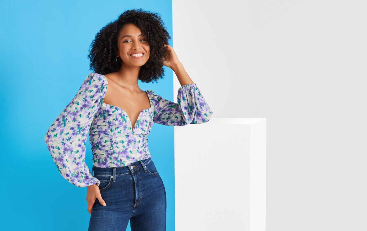 Woman in a floral blouse and jeans posing with one hand in her hair