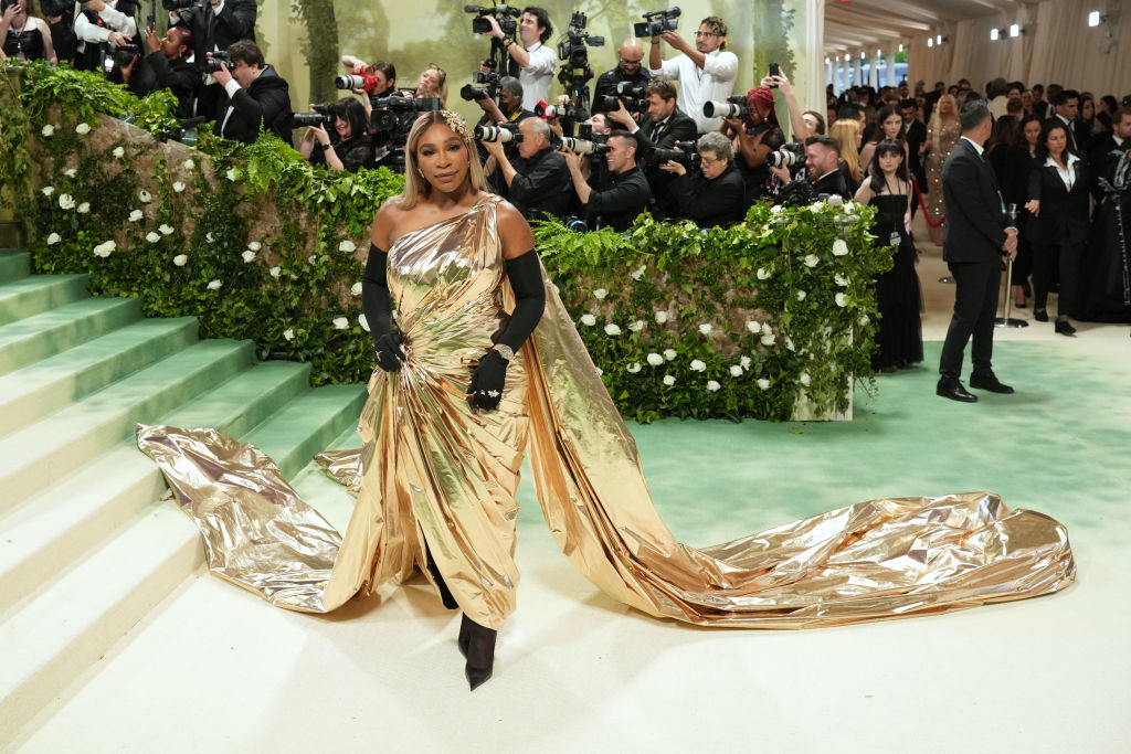 Serena Williams in a flowing gold gown on the Met Gala stairs, cameras flash in the background