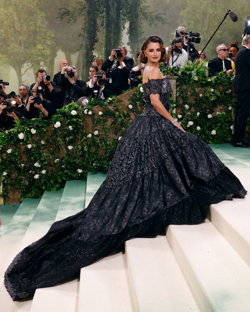 Woman in elaborate off-shoulder gown poses at event with photographers in background