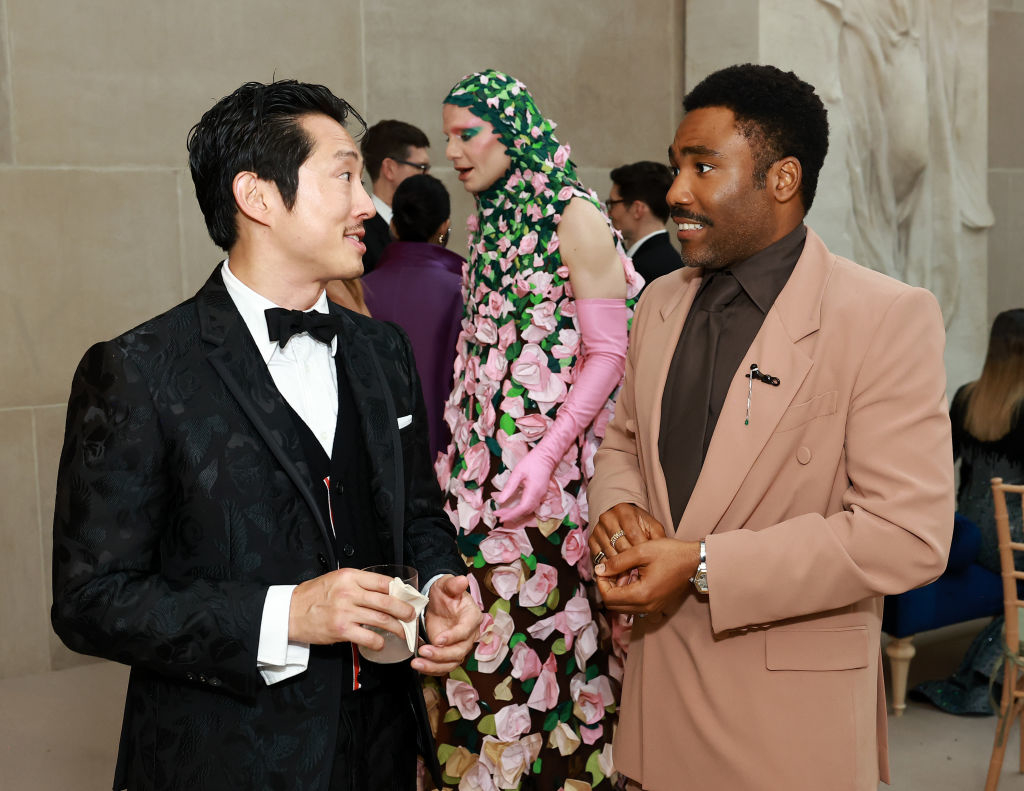 Two men in formal attire conversing, one in a black tuxedo, the other in a tan suit; individual in floral outfit in background
