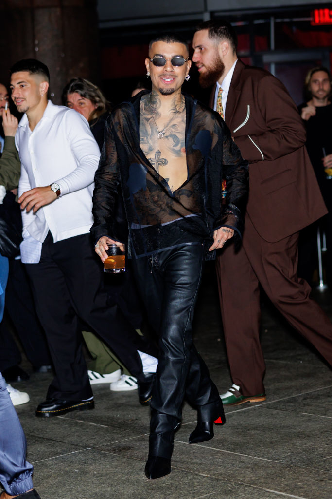 Man with tattoos wearing sheer top and leather pants smiles while walking