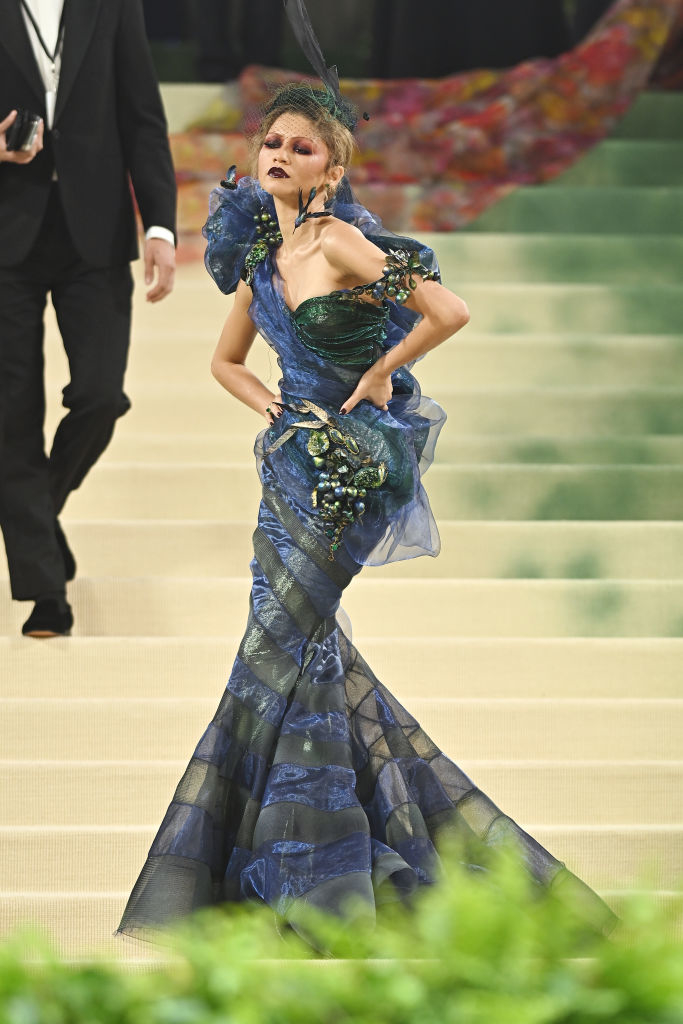A person in an ornate blue gown with ruffled layers and embellishments on a patterned floor