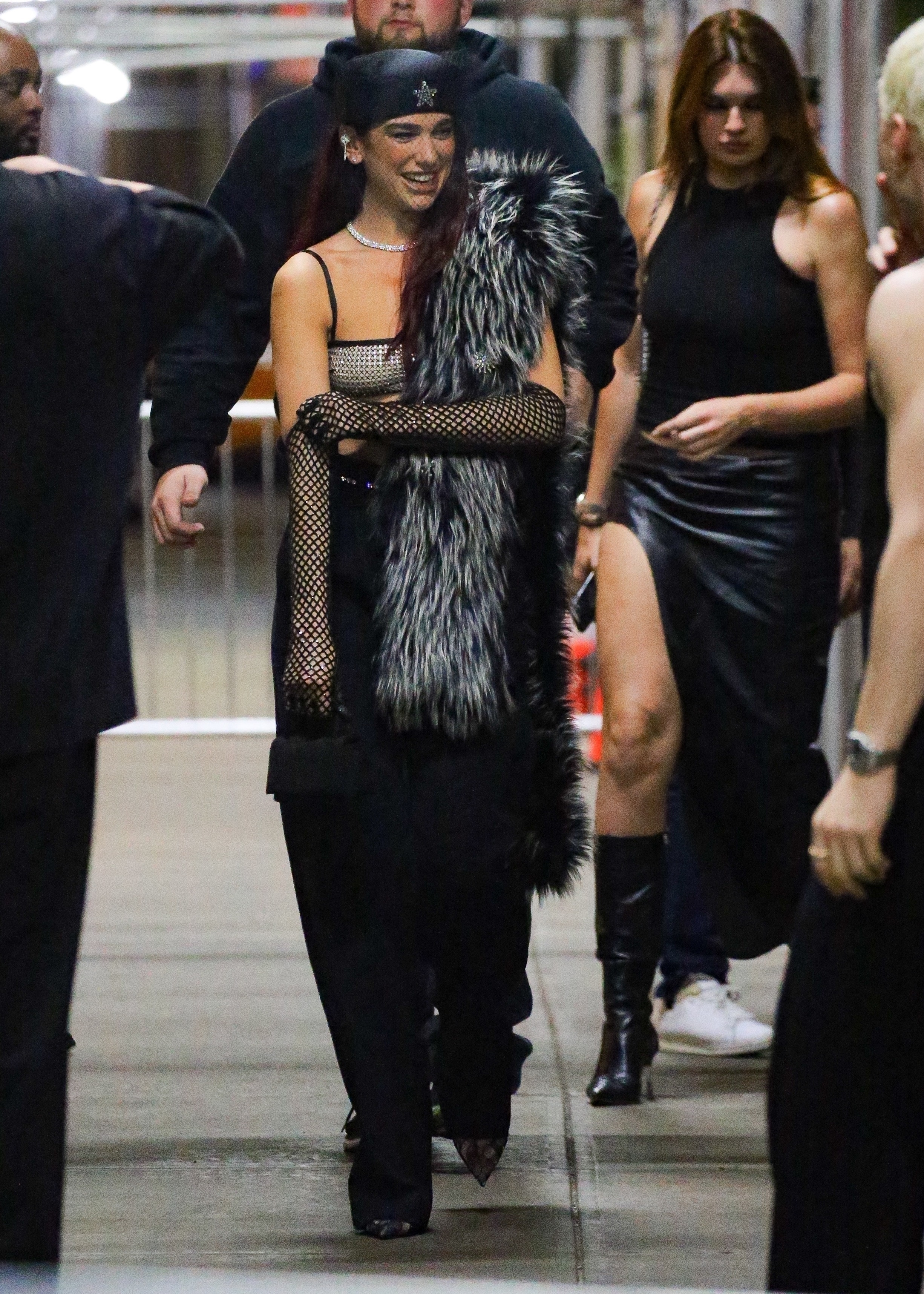 Woman in mesh top with faux fur stole and wide-brimmed hat, walking with others in corridor