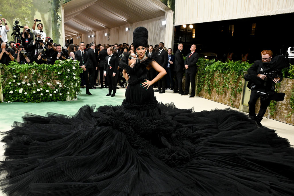 Person in elaborate black gown with voluminous skirt and feathered headpiece on a carpeted event, surrounded by photographers