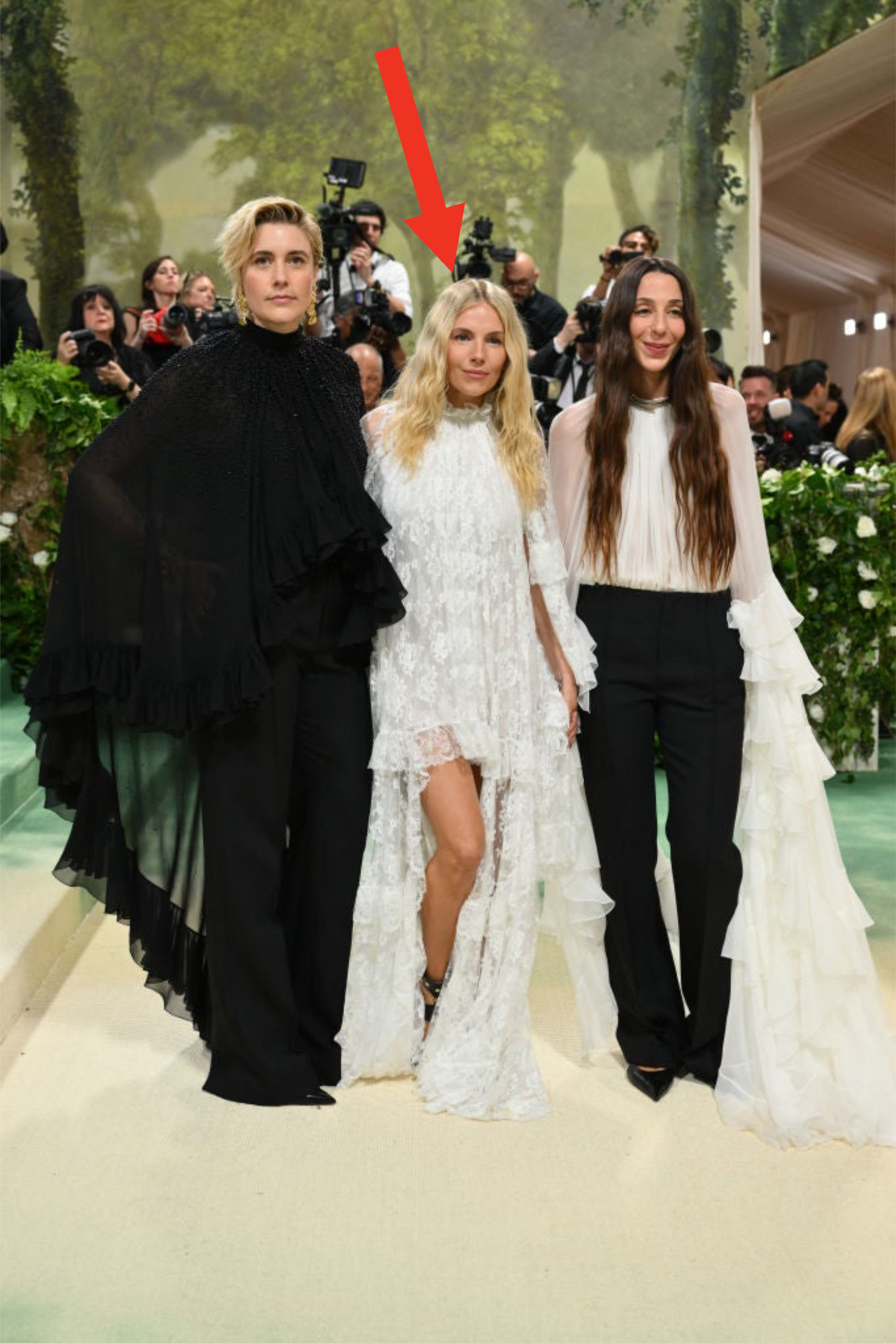 Three people posing at an event; two in dresses with layered designs, one in a suit. They have serious expressions