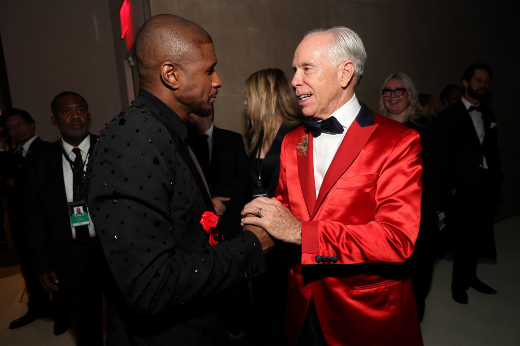 Two men in formal attire shaking hands at an event, one in a suit with decorative motifs, the other in a red satin jacket