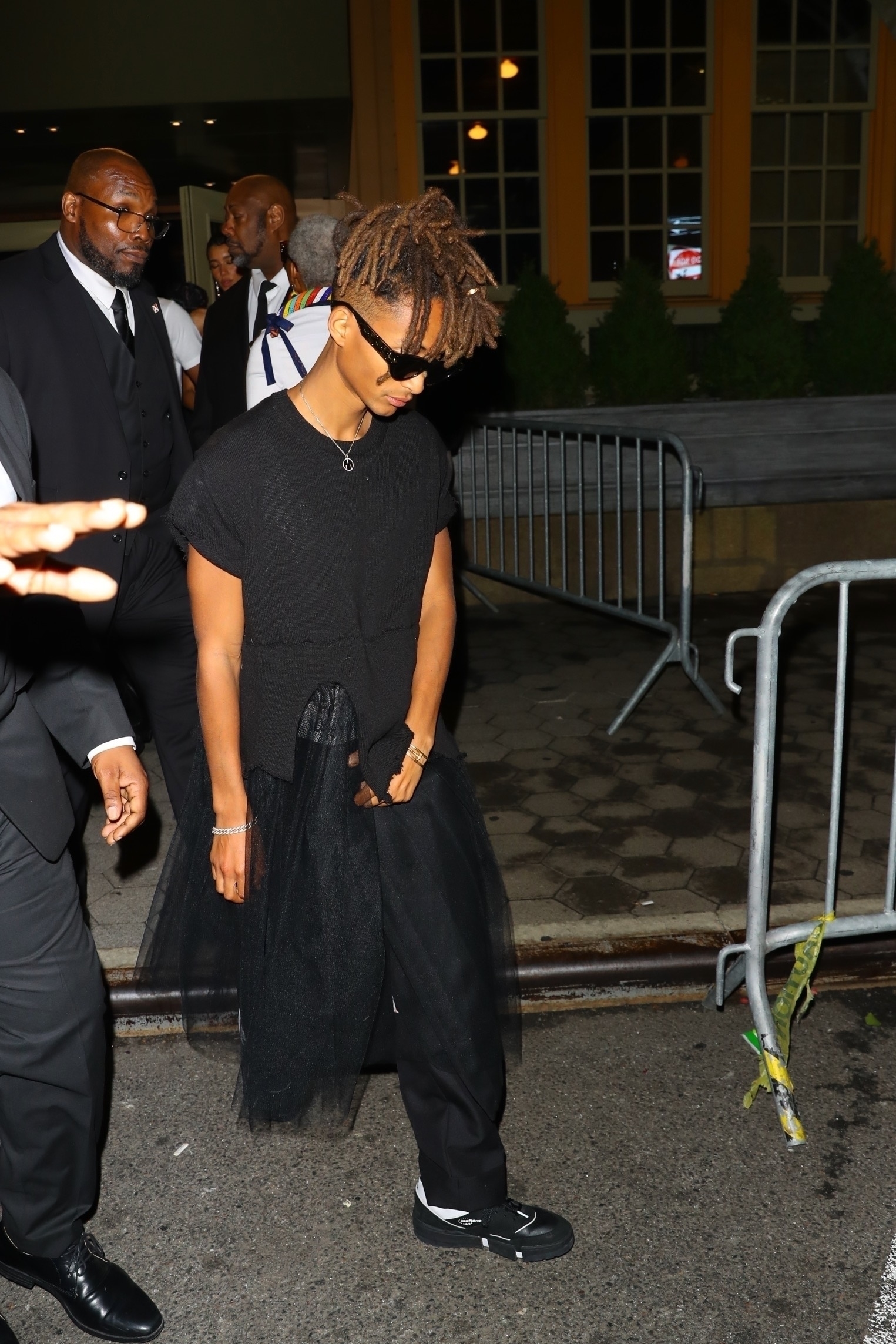 Jaden Smith in a black t-shirt and wide pants with draped detail, sunglasses on, walking past barriers