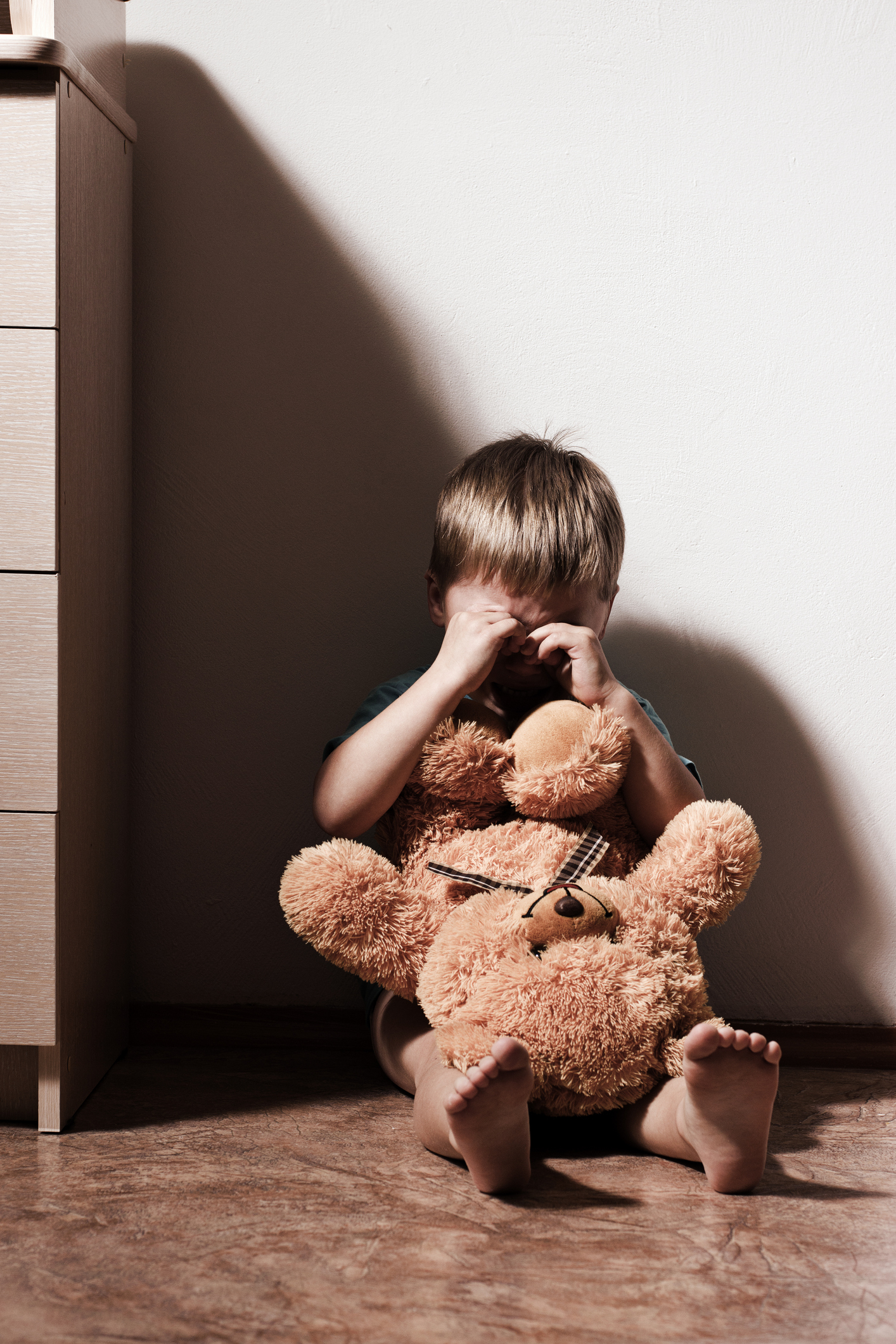 Child sitting on the floor, covering eyes, holding a teddy bear, with a shadow on the wall