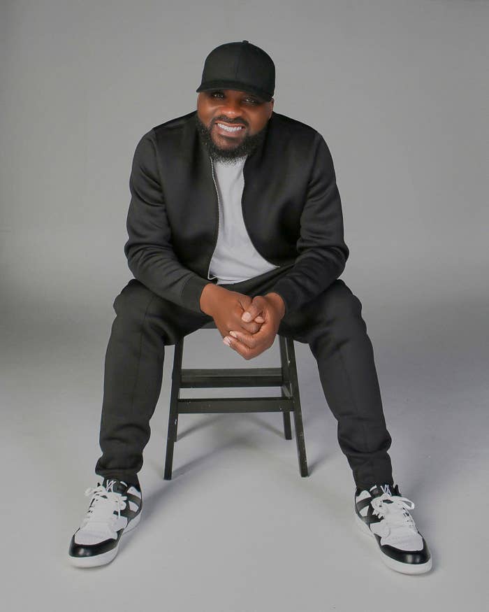 Man seated on a stool, wearing a black tracksuit and sneakers, smiling at the camera