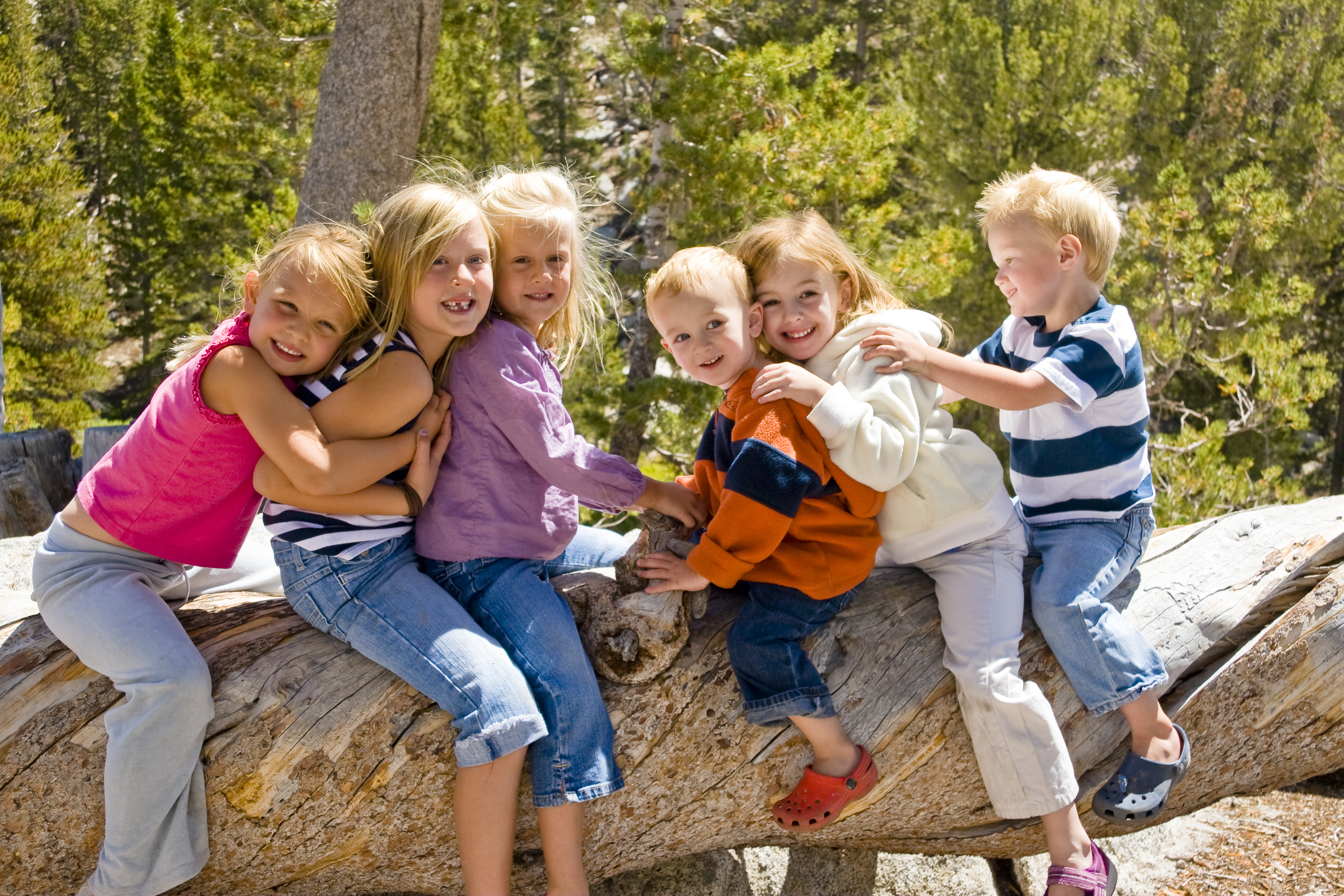 Five children smiling and sitting close together on a large tree log outdoors